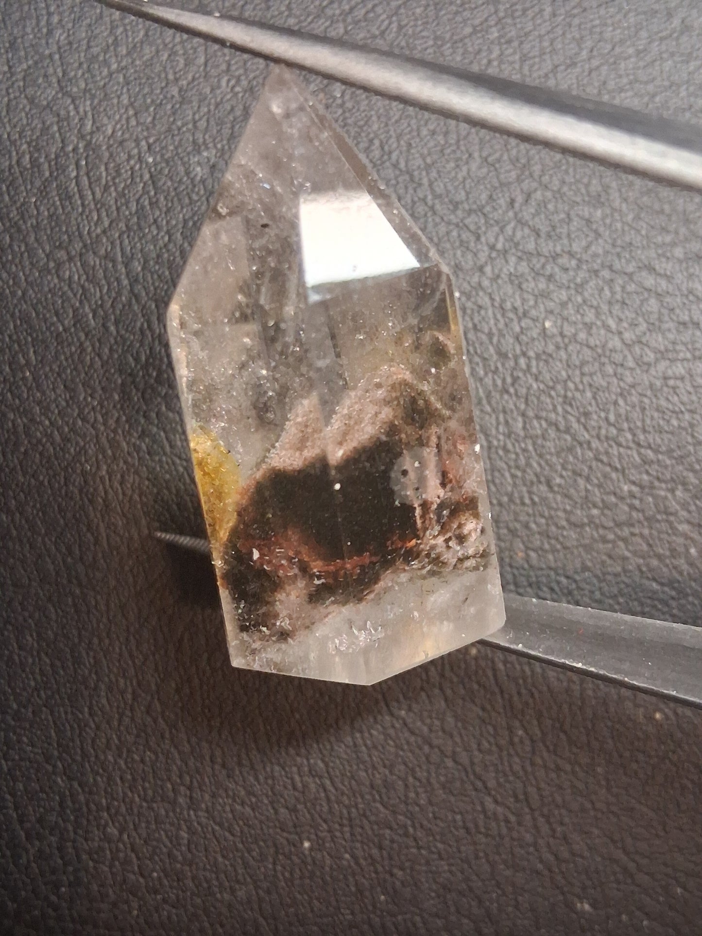 A clear quartz crystal point with internal brown and dark inclusions, held by metal tweezers against a dark textured background.