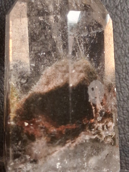 A clear quartz crystal point with a prominent dark brown and black cavity in the center, resting on a dark textured surface.