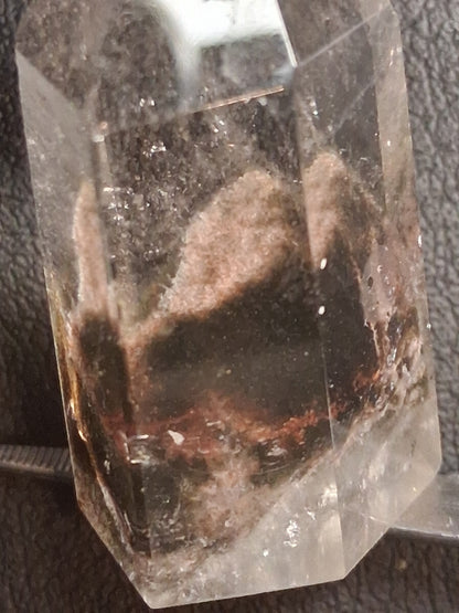 A clear quartz crystal point featuring prominent brown and black inclusions, photographed close-up against a dark textured background.