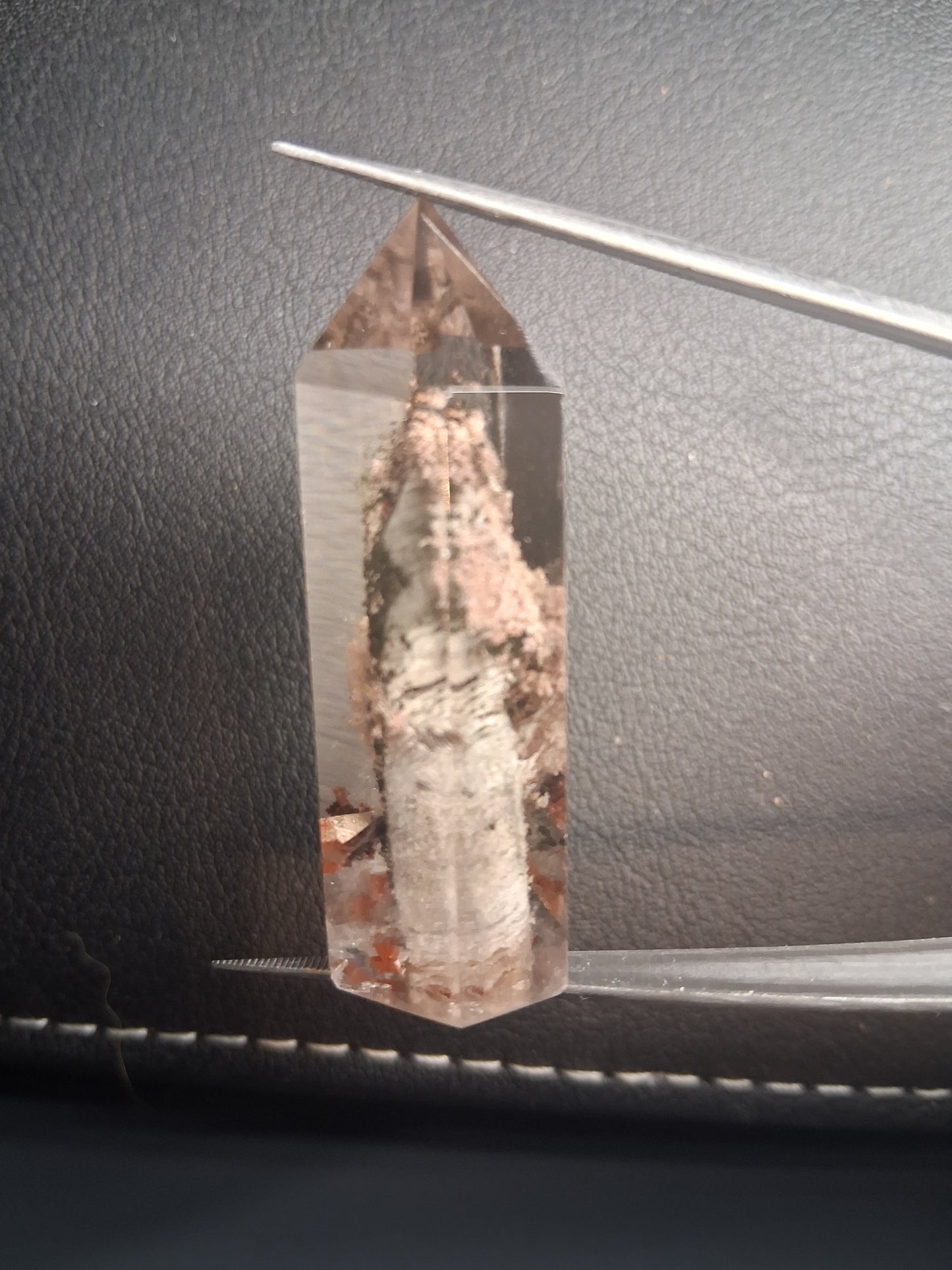 A clear quartz crystal point with rough, earthy brown and beige mineral inclusions, held horizontally by two metal tweezers against a dark textured leather background.