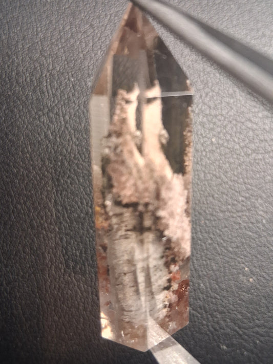 A natural smoky quartz crystal point with internal inclusions, held horizontally by tweezers against a dark textured leather background.