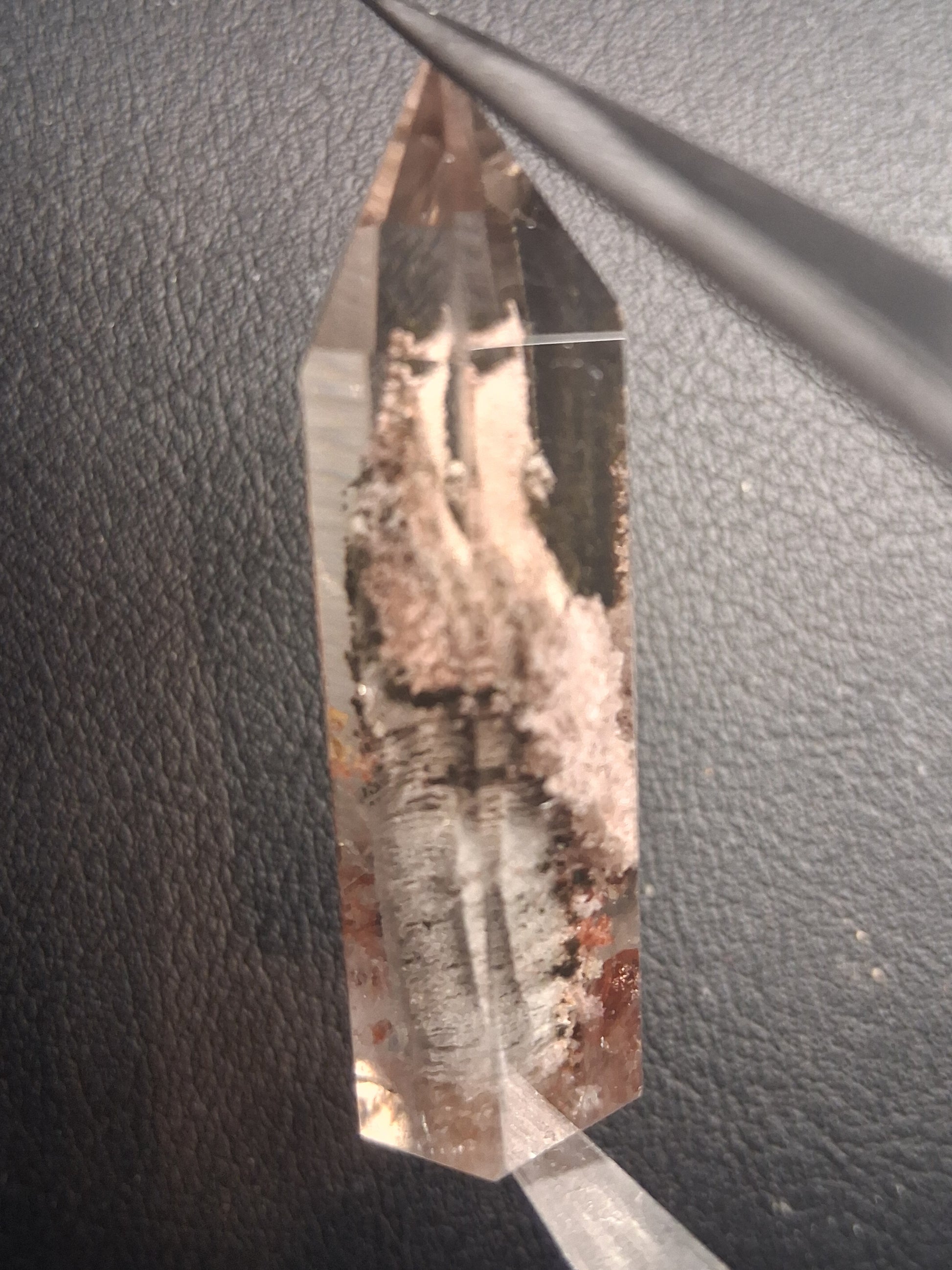 A natural clear quartz crystal point with prominent brown and white matrix inclusions, held by tweezers against a dark textured background.