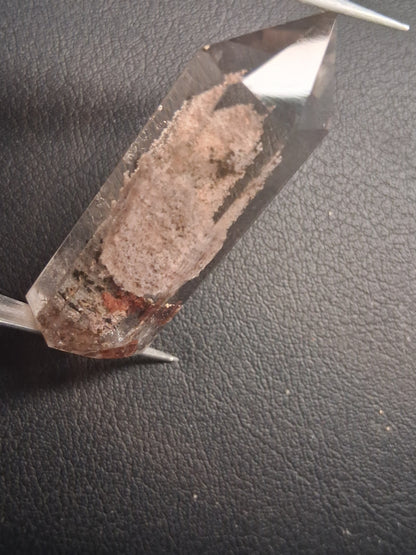 A clear quartz crystal point with a brown and gray matrix inlay, held by metal tweezers against a dark textured background.
