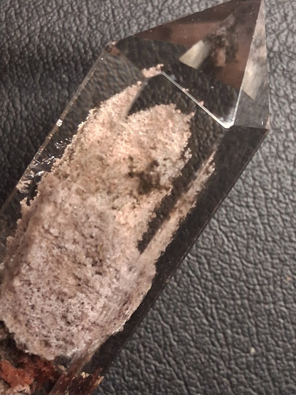 A clear quartz crystal point showing a prominent pinkish-beige and brown mineral inlay, photographed close-up on a textured black surface.