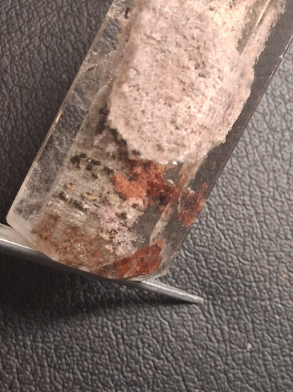 A close-up of a clear cylindrical glass or acrylic tube containing a pale pinkish-beige textured material with some reddish-brown and dark speckled areas, resting on a dark textured surface.