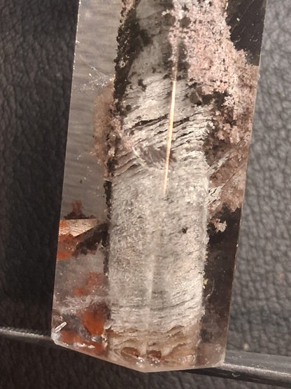 A close-up of a clear quartz crystal point showing natural inclusions, including a prominent white streak, dark brown areas, and small orange mineral deposits, resting on a dark textured surface.
