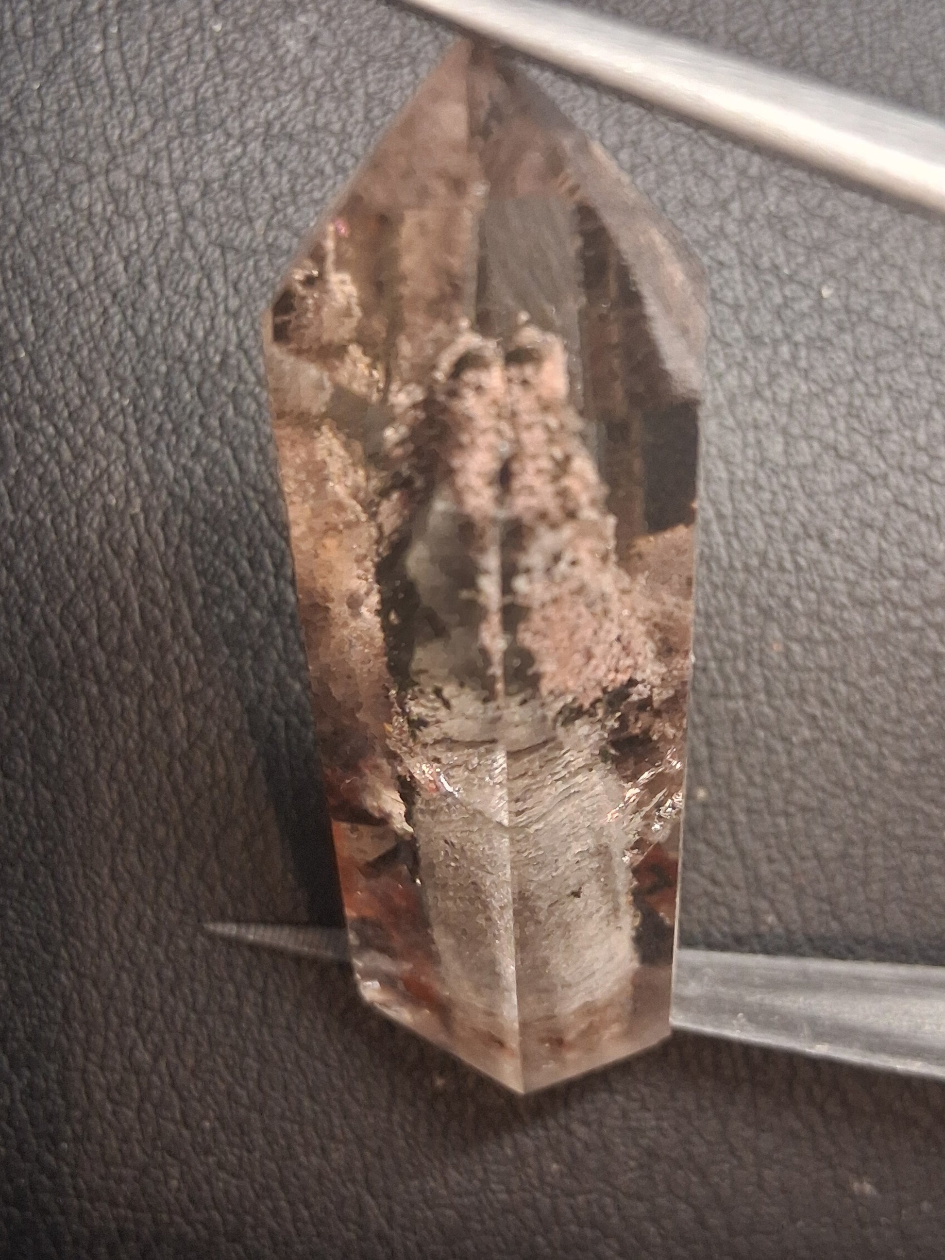 A natural smoky quartz crystal point held horizontally by metal tweezers against a dark textured leather background.