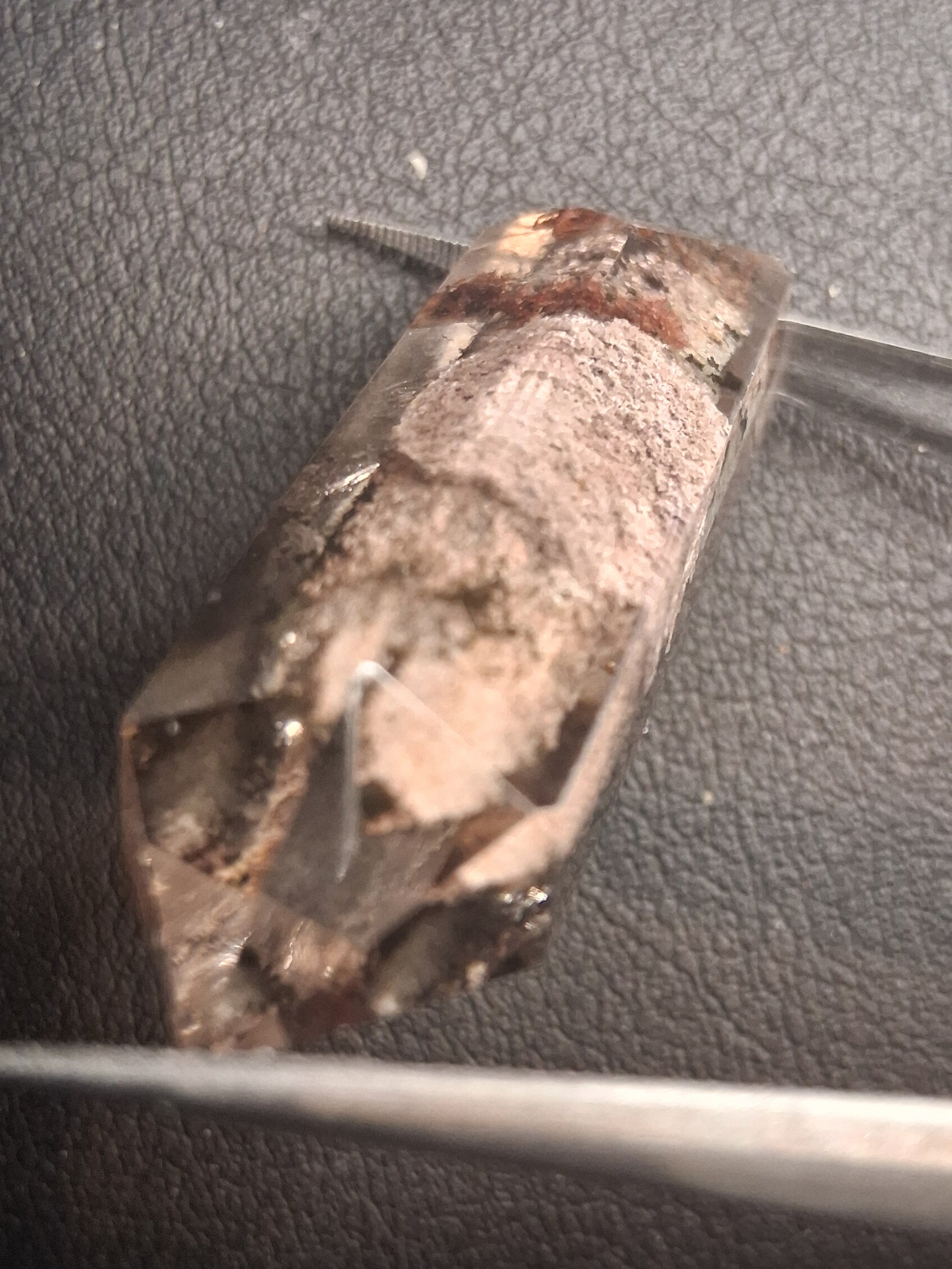 A rough, raw crystal specimen with natural facets and earthy brown and beige tones, held in place by metal tweezers on a dark textured surface.