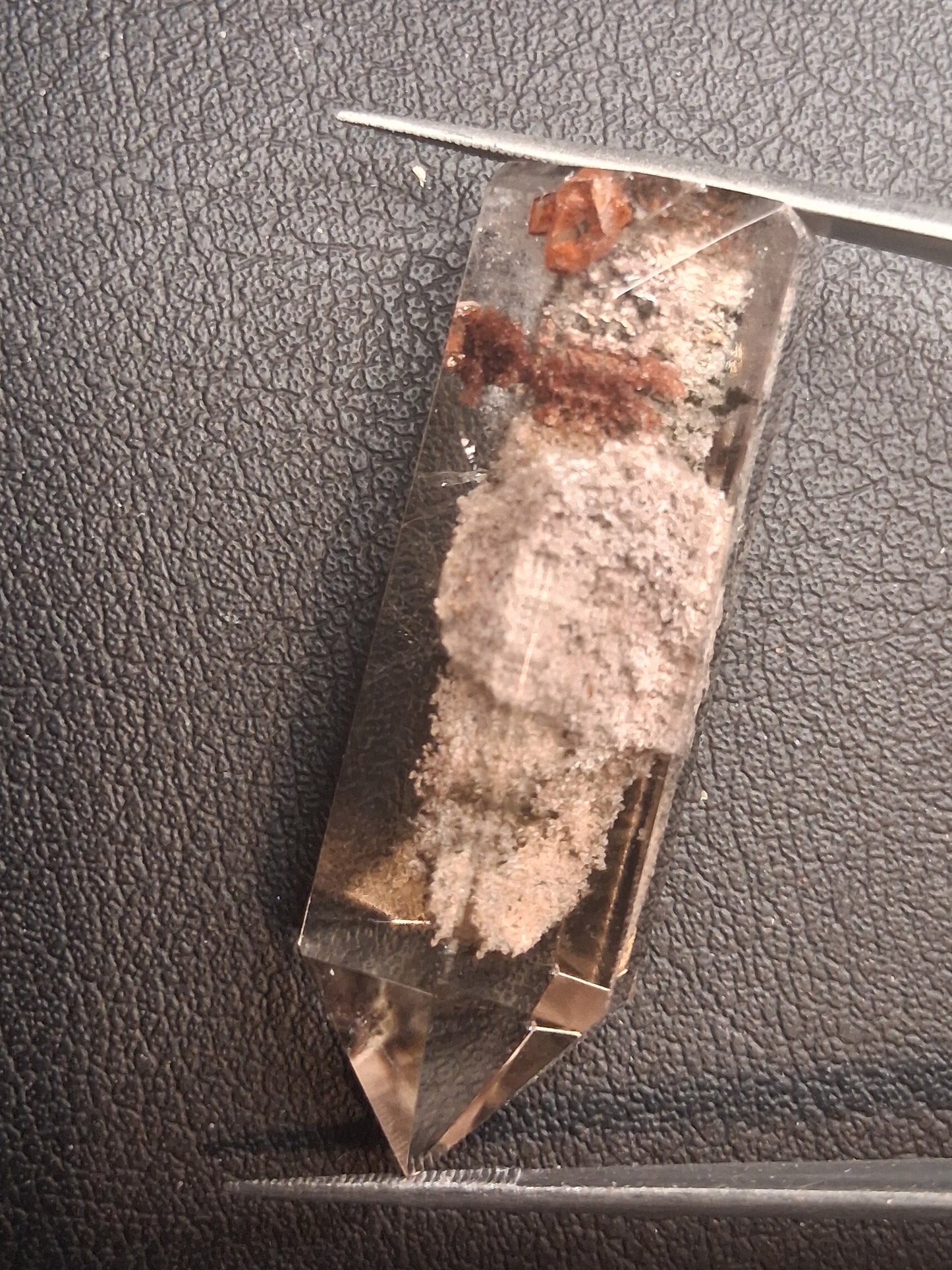 A clear quartz crystal point with internal inclusions, shown on a textured black surface with tweezers visible at the corners for scale.