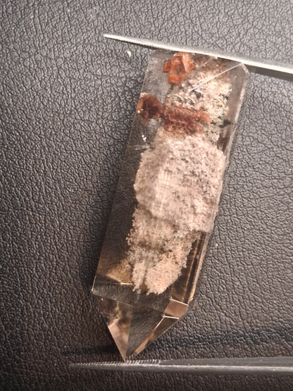 A clear quartz crystal point with internal inclusions, shown on a textured black surface with tweezers visible at the corners for scale.