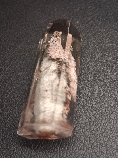 A clear cylindrical glass or acrylic tube with a rounded end, containing a white and pinkish textured substance, photographed on a dark textured surface.