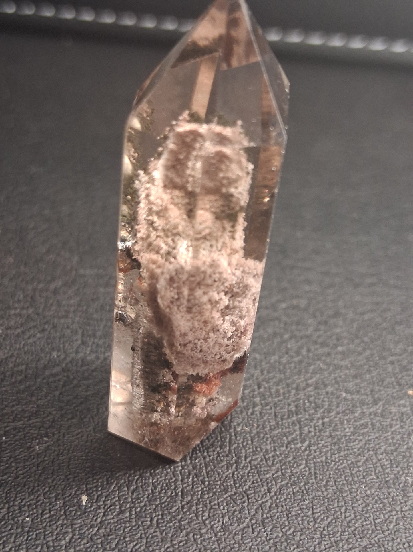 A natural clear quartz crystal point with visible internal inclusions and a rough, dusty center, resting on a dark textured surface.