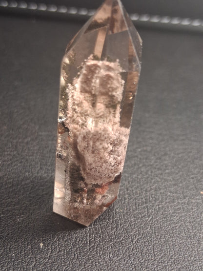 A natural clear quartz crystal point with visible internal inclusions and a rough, dusty center, resting on a dark textured surface.